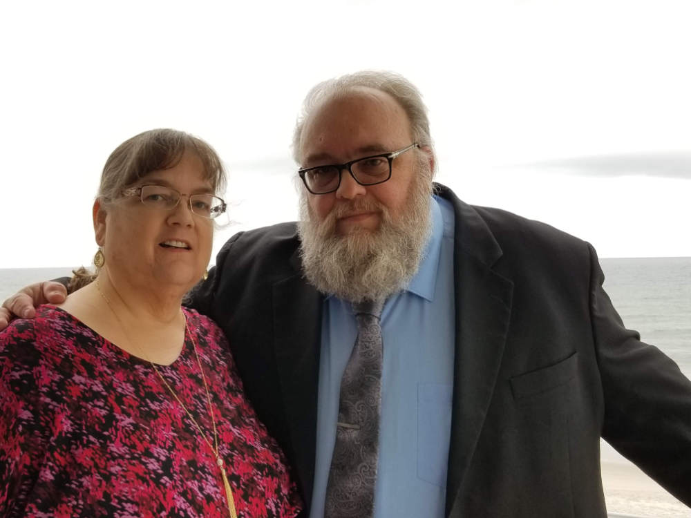 A couple, both well-dressed, appear in the foreground while a pale background of the sea is behind them. The woman on the left is in a flowery red dress, and the man on the right is in a black suit jacket with a light blue shirt and ornate grey tie.
